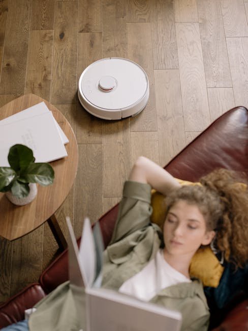 Woman reading on sofa with robot vacuum operating on wooden floor.